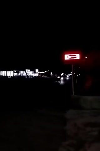 The video shows a dark, rural road at night. A red sign with white text, possibly indicating a destination or a business, is visible on the right side of the road. Several vehicles with their headlights on are traveling down the road. The headlights create streaks of light in the darkness. Buildings with lights on are visible in the background on the left side of the road. The overall impression is of a quiet, dimly lit road with some activity.