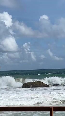 A video shows a choppy ocean with waves crashing against a large rock. The sky is blue with scattered clouds. In the foreground, a wooden railing is visible. The scene appears to be a coastal landscape.