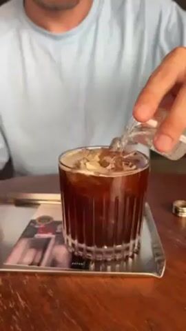 A man sitting at a wooden table pours a clear liquid from a small glass into a larger glass. The larger glass is filled with dark liquid and ice cubes. It rests on a silver tray on top of a brochure. The man is wearing a light blue shirt. The background is blurred.