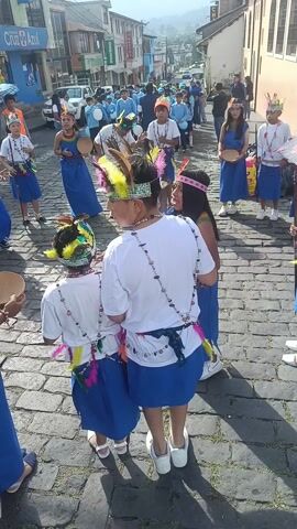 A procession of children and adults moves along a cobblestone street. They are dressed in traditional clothing: royal blue dresses and skirts, white t-shirts, and decorative headdresses and necklaces with colorful feathers and beads. Many hold small, shallow, wooden bowls or thin wooden staves. A few carry produce: a bundle of plantains or a bunch of what appear to be small yellow fruits. Most participants look straight ahead, but some are glancing at each other. The group is passing buildings on either side of the street. A pharmacy is visible in the background. There are spectators watching the procession.  The day is bright.