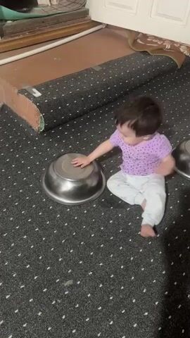 A baby wearing a purple polka-dot shirt and white pants is on a dark gray carpet with a white dot pattern. The baby is interacting with a large metal bowl, pushing it and then stepping on it. Another metal bowl is visible to the right of the frame. In the background, a rolled-up carpet or mat is visible against a wall and a door. The baby's actions suggest exploration and play.