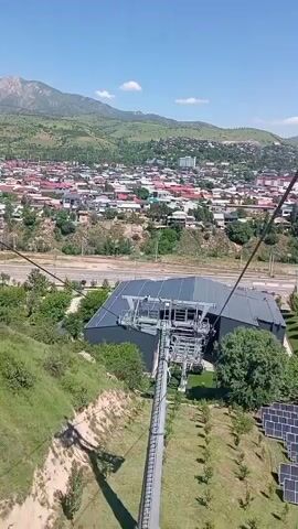 The video is a point-of-view shot from a cable car moving over a cityscape. Initially, the view is dominated by the cable car's structure, the station below, and a solar panel array on a grassy slope. As the cable car ascends, the perspective broadens, revealing a cityscape nestled against a backdrop of rolling green hills and mountains in the distance under a clear blue sky. A river or lake is visible, along with a highway and a large parking lot with parked vehicles.