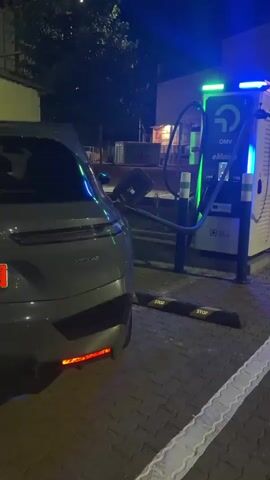 The video shows the rear of a dark-colored electric car parked at a charging station at night. The car's taillights are illuminated, cycling between red and a brighter, almost pinkish-red. A charging cable is connected from the charging station to the car. The charging station is tall and white with blue and green lights and signage that reads "OMV" and "eMotion". The ground is paved with cobblestones and there are parking curb stops in front of the car. Trees and some buildings are visible in the background. The lighting is dim, with the primary light sources being the car's taillights and the charging station's lights.
