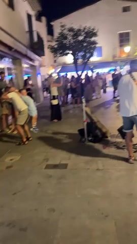 A group of people are gathered in a street at night. In the foreground, a man wearing a white t-shirt and denim shorts is playing a guitar and singing. A small speaker and a water bottle are on the ground near him. To the left, two young boys are leaning against a wall. Further back, a crowd of people are standing and watching the performer. Some people are dancing and others are conversing. Buildings with lights on line the street, and trees are visible. The ground is paved with cobblestones.