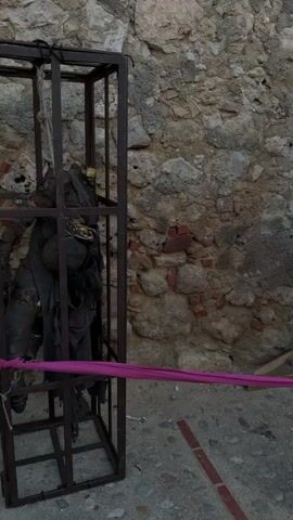 The video shows a small courtyard with historical artifacts related to torture. The scene is staged against an old stone wall. On the left, a metal cage stands containing a dark, possibly skeletal figure adorned with what appears to be a crown. Next to it, a wooden sign titled "LAS JAULAS COLGANTES" (The Hanging Cages) describes the history and context of the cages. Further to the right, another sign, titled "LA SILLA DEL INTERRO" (The Chair of Interrogation), stands near a wooden torture device with sharp spikes. In front of the device, there is a purple mat with various implements, including scissors and a fork. The area is partially cordoned off with a pink ribbon, implying these are exhibits. The ground is a mix of gravel and stone tiles with red markings.