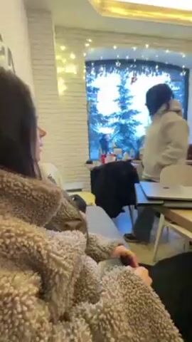 A woman in a tan sherpa jacket sits on a gray sofa in what appears to be a cafe. She has dark hair and is looking off to the right. Behind her, another woman in a gray hoodie and jeans sits at a table with a laptop. Through the large window, snow-covered trees and a few people can be seen outside. There's dance music playing.
