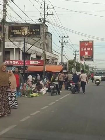 The video shows a street scene with several motorbikes, people, and stalls lining the side of the road. A billboard advertising "RED SCORPION 16" is visible in the background. The road is paved and appears to be in a town or city. Some people are seen riding motorbikes, while others are standing around the stalls, possibly selling or buying goods. The stalls appear to be selling produce, such as fruits and vegetables. The overall impression is of a busy marketplace on a street.