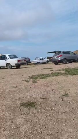 A white car is towing a gray SUV. The cars are on a dirt road. There are other cars in the background. The sky is cloudy.