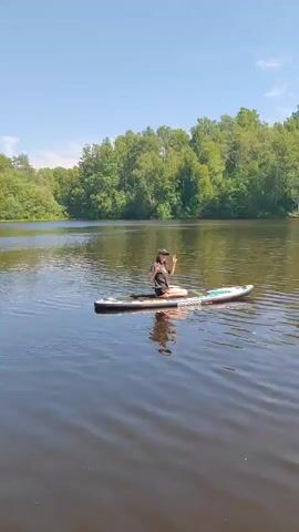A young woman is sitting cross-legged on a paddleboard in the middle of a lake. She is wearing a black t-shirt, shorts, and a baseball cap. She is smiling and looking at the camera. The paddleboard is white with colorful designs. The lake is surrounded by trees. The sky is blue and there are some clouds. The water is calm and reflects the trees and the sky. The woman is raising her hands in a peace sign.