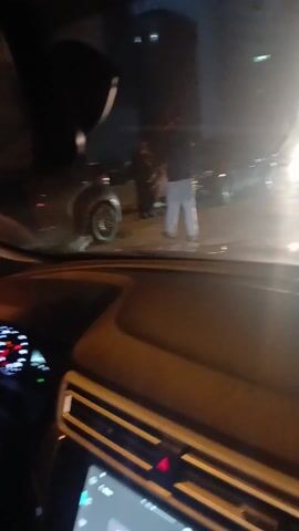 A view from inside a car at night shows people in the street. A man in white pants and a dark jacket is getting into a silver or gray car parked on the street. A woman with a white shopping bag is walking away from the car. Streetlights and buildings are visible in the background. The car's dashboard and steering wheel are visible in the foreground.