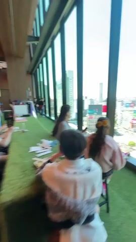 The video shows a view from a high-rise building in Japan, overlooking a busy intersection. Three people are sitting on stools indoors, looking out the large windows. The camera focuses on the cityscape through the windows, showing buildings with various advertisements and signs in Japanese. A large construction site with a crane is visible in the foreground, and many people are crossing the street at the intersection. The sky is clear and blue. The interior space has artificial grass on the floor. The people are not actively interacting with each other.