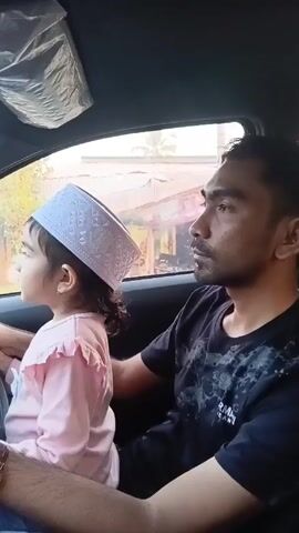 Inside the passenger seat of a car, a young child wearing a white cap sits in the lap of an adult male while he drives. The girl has dark curly hair, a light pink ruffled shirt, and a small earring. She is smiling while gazing out the front window. The man is wearing a black t-shirt with a pattern, and has a beard. His left hand is on the steering wheel with his right arm around the girl in his lap. The exterior shot through the window shows houses and green trees.