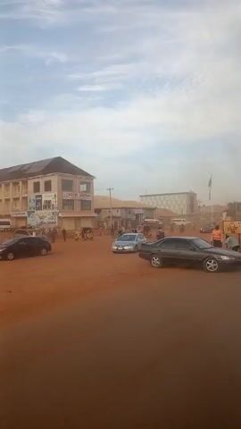 The video captures a street scene with red dirt roads and scattered buildings under a bright sky. Several vehicles, including cars and motorcycles, are present. In the foreground, a dark sedan is parked. To its left, a lighter-colored sedan is visible. Further down the road, a group of individuals are gathered near a yellow signpost, seemingly involved in some activity. Several motorcycles are seen navigating the street. Buildings lining the road appear to be shops or businesses, with one displaying "BEST ELECTRICAL" signage. A large building with a checkered facade stands prominently in the background, and a Nigerian flag is visible on a flagpole. The overall impression is of a bustling, everyday street environment in a town or city.