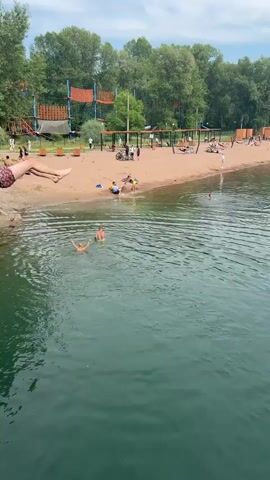 This media file captures a summer day at a sandy beach on a lake. The scene is filled with people enjoying recreational activities. In the foreground, a young man is captured mid-air, leaping from a wooden structure into the clear, green water. The splash he creates upon entering the water is a prominent visual element.

Several other individuals are in the water, swimming and playing. The beach area is populated with people relaxing, some sitting or lying on the sand, while others are gathered around in small groups. Bicycles are visible near the beach, suggesting active use of the area. In the background, a large, multi-level adventure park structure with orange and blue elements stands out against the lush green trees that line the shore. The sky is partly cloudy, indicating fair weather conditions. The overall atmosphere appears to be relaxed and cheerful, with a focus on outdoor recreation. The location cues, such as the type of vegetation and the overall setting, are consistent with a beach environment in Abakan, Russia. There is no visible text in the media file.
