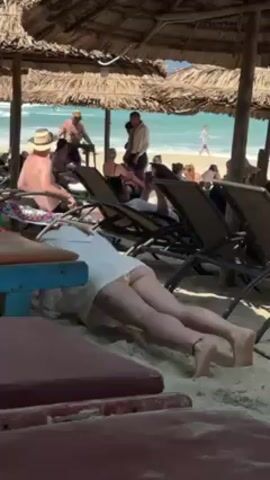 The video depicts a beach scene under a thatched-roof structure, likely a beach hut. In the foreground, a woman in a white top is doing push-ups on a table, exposing her buttocks due to the top's length. Several beach chairs are visible around her, some occupied by people. In the background, the ocean and a crowded beach are visible, with people walking and sunbathing. The overall impression is a casual, sunny beach environment.
