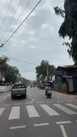 The video shows a road in what appears to be a tropical or subtropical area. A gray pickup truck is driving on the left side of the road. There are other vehicles present, including a green tuk-tuk and a blue tuk-tuk. There are people on a scooter in the foreground, and other pedestrians are visible further down the street. The road is marked with white lines, and there is a crosswalk in the foreground. The area has buildings, trees, and power lines. The sky is overcast.