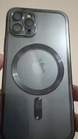 A person is holding a gray cell phone with a protective case in front of a plain, off-white background. The phone has a circular design element on the back with the Apple logo visible through it. There is also a metallic strip below the circle, near the bottom of the phone. The camera lenses are visible in the top left corner of the phone's back. The person's fingers are slightly visible on the left side of the phone, holding it in place. Sounds of a child talking can be heard.