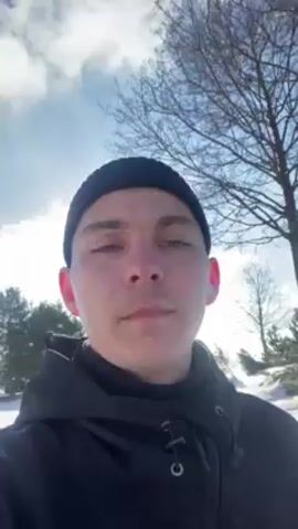 A young man wearing a black beanie and a black jacket is recording a video of himself outside on a bright, sunny winter day. There is snow on the ground and some trees are visible in the background. The sun is shining brightly, creating a glare in the camera. The man is looking directly at the camera with a neutral expression. He is standing on a paved path. The background shows a snowy landscape with trees and some distant structures.