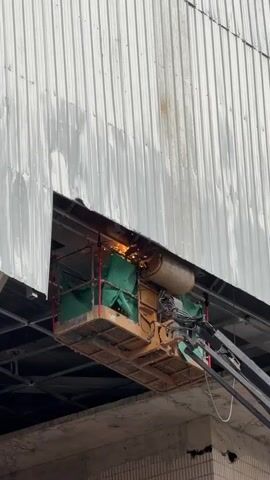 In Qinhuai District, China, a scissor lift is positioned near the side of a large, corrugated metal building. The building's exterior is a pale, reflective white, marked by streaks of rust and water stains. A large, rectangular opening has been cut into the metal siding, revealing the structure beneath.

Within the scissor lift's platform, a worker is engaged in welding. Sparks fly from the welding torch, creating a bright, incandescent glow. The platform is partially covered by green tarps, and a large, cylindrical object, possibly a fuel tank or other piece of equipment, is mounted on the lift. The scene appears to be during daylight hours, though the specific time of day and weather conditions are not clearly discernible.

The overall impression is one of industrial work or repair being carried out at height. The raw, unfinished edges of the cut opening and the dynamic nature of the welding activity suggest ongoing construction or maintenance. The surrounding environment below the lift appears to be a concrete structure with visible brickwork, hinting at an industrial or commercial setting. No visible text is present in the media.