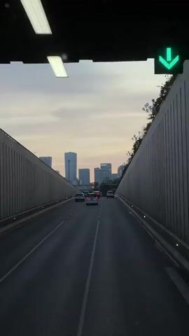 The video shows a street with several cars driving toward the camera.  The street has high walls on either side, and between these walls, buildings are visible in the distance.  There is a twilight sky behind the buildings.  Streetlights are positioned along the top of the walls.