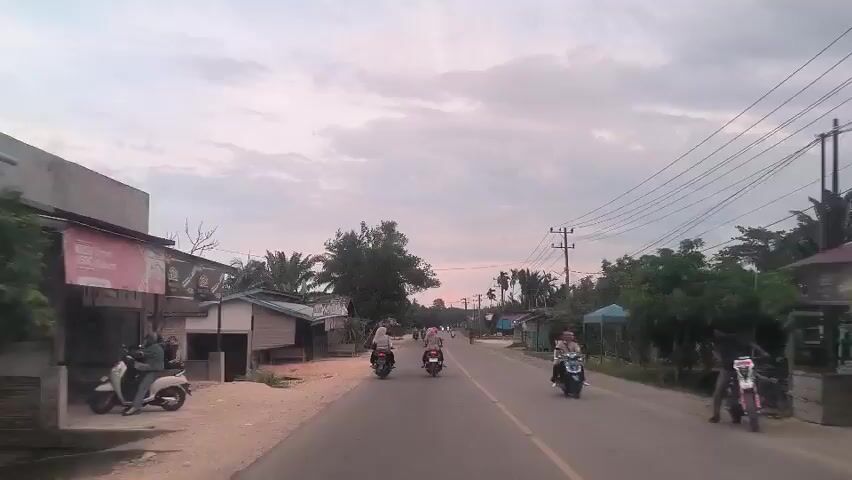 The video captures a drive down a paved road, likely in a rural or semi-rural area of Southeast Asia. The sky is overcast with hues of pink and gray, suggesting either sunrise or sunset. Alongside the road, there are various small buildings and houses, many appearing to be modest in construction. Several utility poles with overhead wires run parallel to the road, adding to the rural atmosphere. A number of people are seen riding motorbikes along the road. The video seems to be taken from the perspective of a vehicle moving along the road.