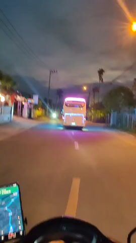 The video shows a first-person perspective from a motorcycle following a white minibus at dusk. The minibus has pink and blue neon lights outlining its shape. Streetlights illuminate the road and nearby buildings. Power lines stretch across the sky. A phone is mounted on the motorcycle's handlebars, displaying a map. The road has a single white line indicating lanes. There are trees and residential buildings on either side of the road. The sky is a mix of dark and light clouds, indicating the transition from day to night.