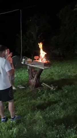 A person is walking around a backyard at night. There is a fire burning in a grill, a swimming pool, and a wooden structure with a television and lights. The person briefly stops to look at the fire. There are other people in the background.