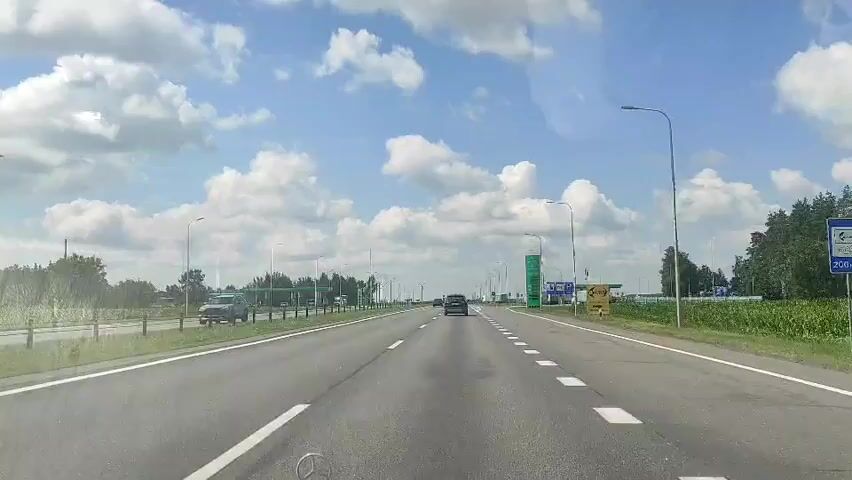 The video shows a view from a vehicle traveling on a highway. The sky is blue with scattered white clouds. On the left side of the road, there are trees and what appears to be a gas station with green pumps. On the right side, there is a cornfield and a tall green sign with writing and numbers, likely indicating fuel prices. There are streetlights along the highway. Several other vehicles are visible on the road ahead, including a car and a truck. The highway has multiple lanes and white dashed lane markings. The overall environment suggests a sunny day on a rural highway.
