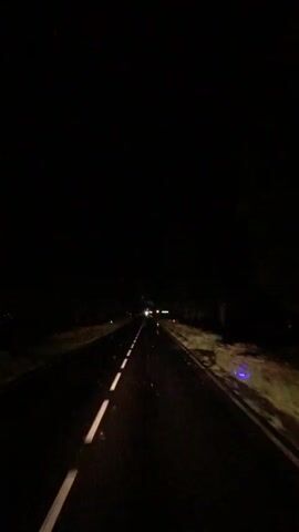 The video shows a dark, rural road at night. The perspective is from inside a vehicle, likely driving. The headlights of other vehicles can be seen in the distance. White lines mark the edge of the road. There are also some small blue lights visible along the side of the road, possibly reflectors or markers. The surrounding environment is dark and appears to be unlit. There are no people or clear identifiable objects other than the road and other vehicles.