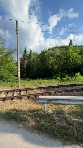 The video captures a first-person perspective of someone riding a scooter on a paved road. The setting is rural, with abundant greenery and trees lining the road. A railway track crosses the road, indicated by the visible rails and a crossing gate. The sky is blue with scattered white clouds, suggesting a pleasant day. The time of day appears to be daytime, likely late morning or afternoon.

The rider is wearing a black t-shirt and appears to be in motion, turning the handlebars of the scooter. The scooter itself is black and has a rearview mirror. Visible text on the scooter includes warning labels with symbols and text that appears to be in a non-Latin alphabet, possibly related to regulations or product information. There are also railroad crossing signs visible, one of which has markings that might be numbers or symbols. A small, weathered building with a sloped roof is visible near the railway tracks. The overall mood seems relaxed and casual, indicating a leisurely ride through the countryside. The location is identified as Boltata, Bulgaria.