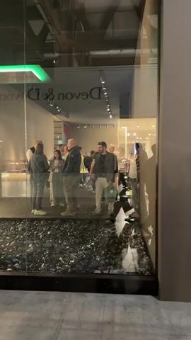 The video opens with a view into a brightly lit interior space through a large glass window or partition. Reflected on the glass, the words "Devon & Devon" are visible in reverse, suggesting a brand name for the establishment. Inside, numerous people, a mix of men and women of various ages, are standing around in small groups, conversing and observing their surroundings. The setting appears to be a modern showroom or exhibition, possibly displaying bathroom fixtures or home furnishings, as hinted by what look like sinks or vanities in the background.

In the foreground, outside the glass, there's a decorative feature resembling a shallow water basin filled with dark, possibly black, polished stones or pebbles. A striking green neon-like light strip is visible on the ceiling above the interior space.

The camera then pans slightly to the right, moving past a plain, solid gray wall. As it continues to pan, it reveals another large display wall with a vibrant yellow and blue graphic, featuring some indistinct text. Next to this wall, a small white sign on a stand clearly displays the alphanumeric code "C38." The overall atmosphere suggests an event, possibly an industry exhibition or showroom opening, given the number of attendees and the curated displays in Rho, Italy.
