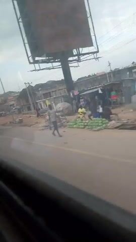 The video shows a street scene with several buildings under construction or renovation. A motorcycle is visible on the road, and several people are walking around. The ground appears to be unpaved and dusty in some areas. The sky is overcast. The perspective suggests the video is being filmed from a vehicle.
