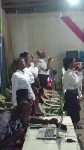 A group of about seven women, dressed in white shirts and black skirts, are standing together and singing into microphones in an indoor setting. One woman has a patterned head covering. Behind them, a row of chairs is visible, with a red drum set partially visible behind the center singer. In the foreground, a man sits at a table, working on a laptop. The room appears to be a simple, possibly church-related, space with a plain wall and a red cloth decoration hanging in the background. The lighting is provided by a single bright ceiling lamp.