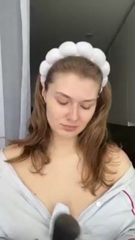 A woman is applying makeup in front of a window with sheer curtains. She has long, light brown hair, fair skin, and is wearing a white, cloud-shaped headband and a light blue and white striped pajama top. She holds a large, black makeup brush in her right hand and applies makeup to her face. She looks directly at the camera with a slight wink. In the background, there is a glimpse of what appears to be a room with light-colored cabinets. The lighting is soft and natural.