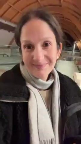 A woman in a black fleece jacket and gray scarf smiles at the camera. She is in a large, well-lit room with other people. Some are seated at tables. The room has a wooden frame structure.