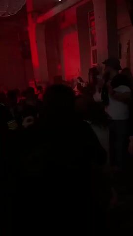 The video shows an indoor space, dimly lit with a strong red hue from overhead lights. A large crowd of people, primarily young adults, occupies the room, standing and moving. The camera is handheld and moves slightly, positioned within the crowd. Many individuals are partially obscured by darkness or other people. A prominent person in a black and yellow sports jersey with "PICKENS 14" on the back is visible moving through the frame. Several women with long hair and light-colored tops are seen closer to the camera. In the background, there are architectural features like pillars and a stage or elevated platform with more red lighting, where indistinct figures are visible. A white, sculptural light fixture hangs from the ceiling. Music is audible in the background.
