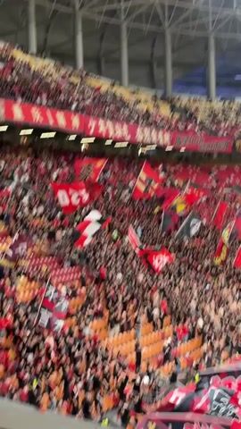 The video shows a packed stadium full of fans waving red and black flags. The stands are primarily red and orange, with a sea of fans dressed in dark clothing, creating a strong contrast. The flags, varying in design, add to the energetic atmosphere. Banners with Chinese characters hang above the crowd. The stadium's architecture includes visible support structures.