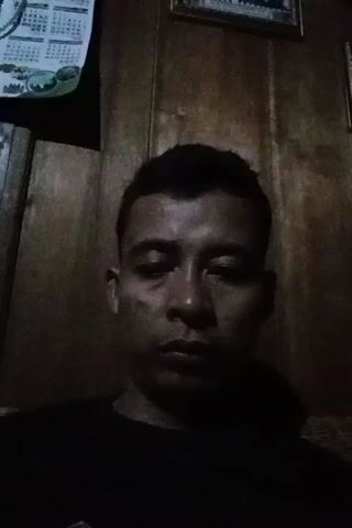 The video shows a close-up, head-on, waist-up shot of a man. He has short dark hair and is wearing a black t-shirt. The background appears to be a wooden wall with a calendar and a framed picture hanging on it. The lighting is dim and casts shadows on the man's face. The man's expression is neutral, and he does not move throughout the short clip.