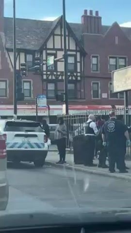 The video shows a street scene with multiple people gathered on a sidewalk. A police car with flashing lights is parked nearby. Several individuals appear to be interacting with law enforcement officers, some of whom are wearing vests and surgical gloves. The people are standing near a trash can and a fence. A building with a Tudor-style façade is visible in the background. The overall atmosphere suggests an active police investigation or response to an incident.