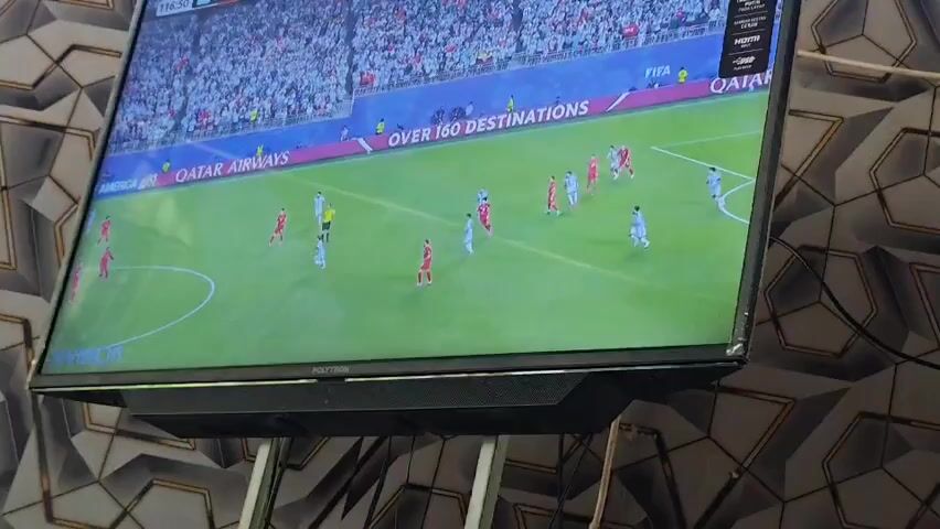 A television screen displays a soccer match in a stadium. The field is green, with white lines indicating boundaries and markings. Several players in red and white jerseys are visible on the field, engaged in the game. Spectators are seated in the stands, mostly wearing white. Advertising boards line the perimeter of the field, featuring logos and text, including "Qatar Airways" and "FIFA World Cup 2022". The TV screen is mounted on a wall with a distinctive patterned wallpaper.