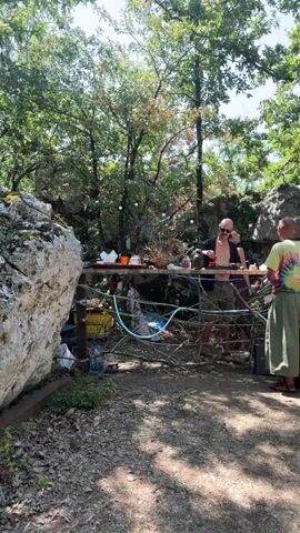 A video shows a group of people outdoors in a wooded area with large rocks and trees. In the foreground, a person wearing a tie-dye shirt and a long green skirt stands facing the camera. Behind them, a table is set up with various items, including bottles, cups, and what appears to be food or drinks. Two other people are visible behind the table, interacting with the items on it. One person, wearing sunglasses and an open shirt, is leaning over the table. The other person, with their back to the camera, is also near the table. The overall atmosphere suggests a casual gathering or event in a natural setting.