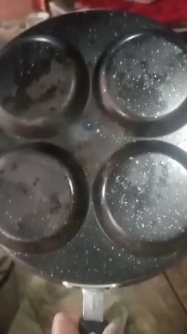 A person's hand holds a black frying pan with four round indentations. The pan is speckled with white. The indentations appear to be lightly oiled. The person is preparing to make mini martabak.