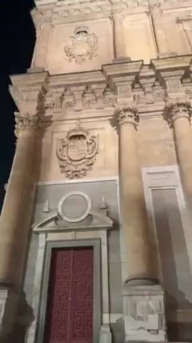 At night, a low angle shot reveals a large building with columns and decorative details, likely a church. The building is lit up, and a red door is visible at the bottom. The camera pans upwards to show the top of the structure. People are present in the background, but they're not the focus of the shot.