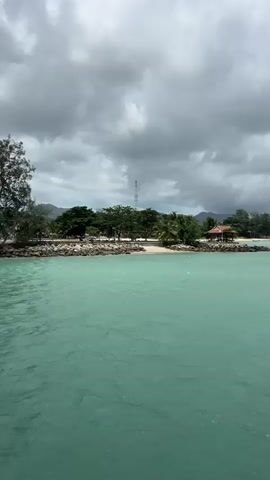The video shows a cloudy day at the beach. The water is turquoise and there is a small building on the beach with some trees around it. There is a tower visible in the distance. The camera pans across the ocean to show more of the water and the horizon. The sky is overcast with dark clouds. There are a few small sailboats on the water.