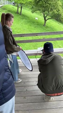 A small film crew is shooting footage of a table with several small beakers or cups on it outdoors. The table is on a wooden porch overlooking a grassy hill. A woman in a green jacket and jeans holds a light reflector. A man in a green jacket and a baseball cap crouches down to adjust the camera. Another person, in a dark blue puffer jacket, stands to the side, partially obscuring the view. In the background, a small white animal stands on the green hill.