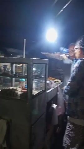 A low-light, handheld video shows an outdoor food stall at night. A glass-enclosed display case, likely for food items, is visible. A person is standing near the stall, partially obscured by shadow and motion blur. In the background, a bright light shines, illuminating a portion of the scene. Further in the background, a motorcycle passes by, and there are indications of other buildings or structures. The overall environment appears to be a street or market setting.