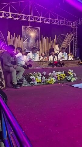 A group of musicians perform on a stage illuminated by purple lights. They are playing traditional Indonesian instruments, likely part of a gamelan ensemble. The stage is decorated with bamboo poles and floral arrangements. Two individuals sit on the left side of the stage, observing the performance. The performance appears to be taking place outdoors or in a large open space, with a truss system visible above the stage. The floor in front of the stage is covered with a magenta carpet.