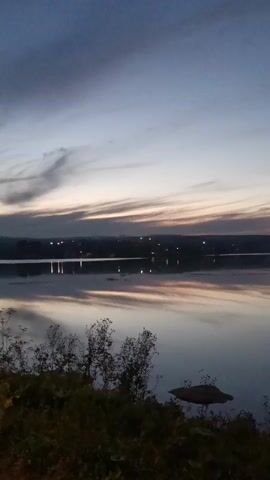 The video shows a serene lake at dusk. The sky is painted with soft hues of blue, gray, and pink, with wispy clouds streaking across the horizon. The water is calm and reflects the sky, creating a mirror-like effect. A distant shoreline is visible, dotted with faint lights that suggest a town or village. In the foreground, silhouetted trees and foliage add a touch of depth to the scene. The overall atmosphere is peaceful and tranquil, evoking a sense of quiet contemplation.