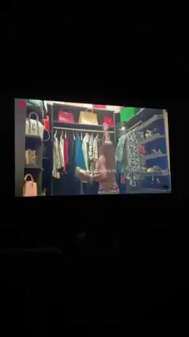 A video shows a woman standing in her closet. She is wearing a striped, form-fitting dress. Her closet has multiple shelves containing handbags and shoes. There is also a hanging rack full of clothes. The walls and shelves are dark. The video is dark as well, but the closet is brightly lit. At the bottom of the screen, a bit of a person is seen, sitting, presumably, watching the video. The bottom of the video has text, BIMBO ADEMOYE TV.
