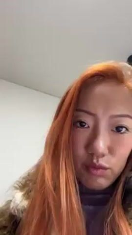 The video shows a woman with long orange hair, wearing a fur jacket and a purple turtleneck shirt. She is holding up her right hand, showing off her extra-long fingernails. The nails are clear with silver and gray accents, and some have rhinestone decorations. She flexes her hand into a claw shape. The background is a plain white wall. There is faint chatter in the background.